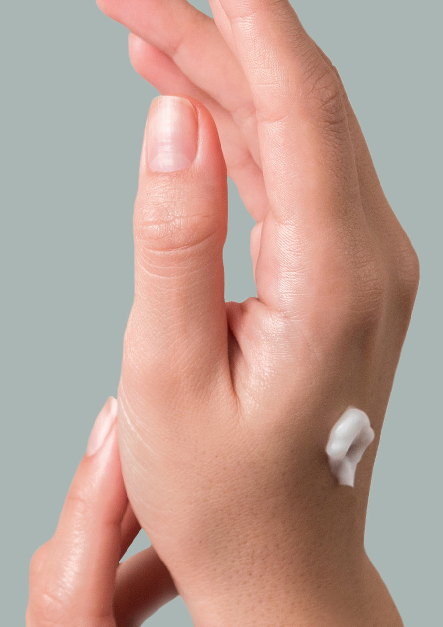 Hand with a dab of white hand cream on the back against a sage grey background
