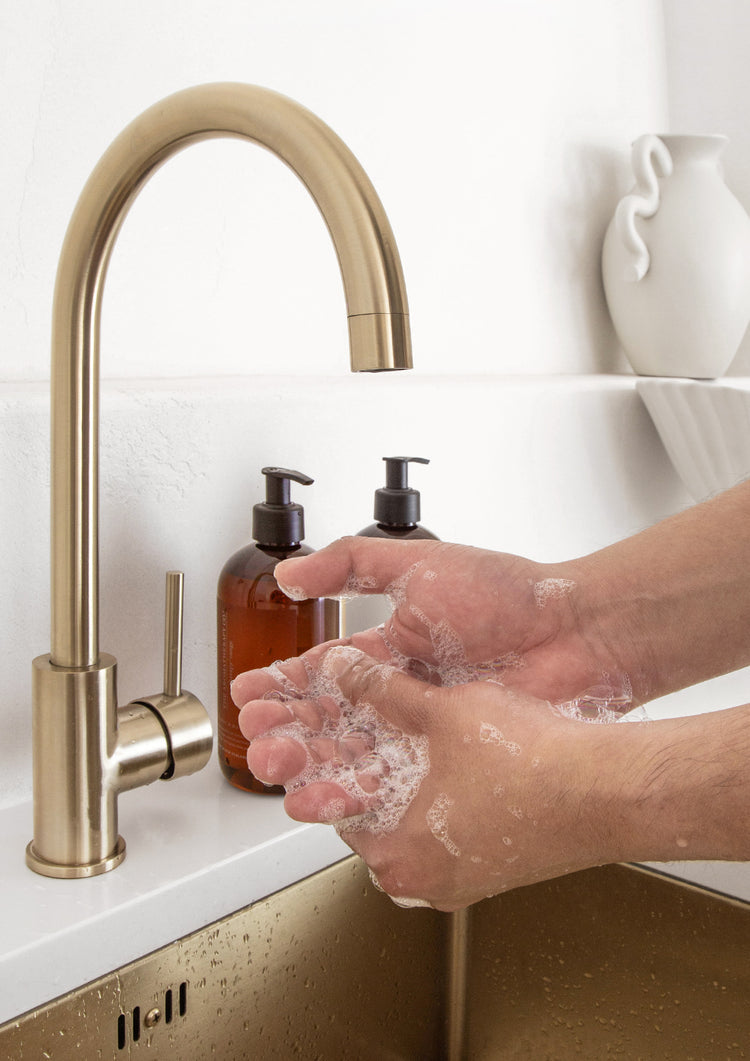 Hands lathering under a brass gooseneck tap with two Therapy bottles on the bench