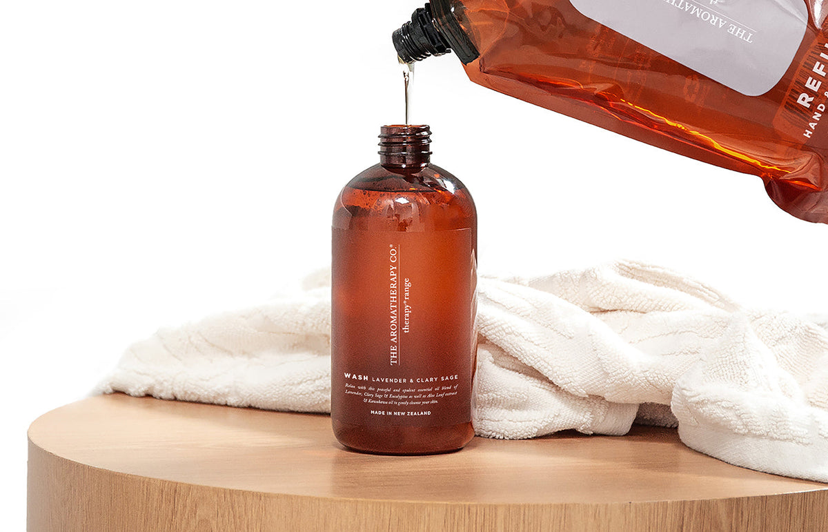 Refill pouch being poured into an amber Therapy bottle on a round timber stool with a white towel