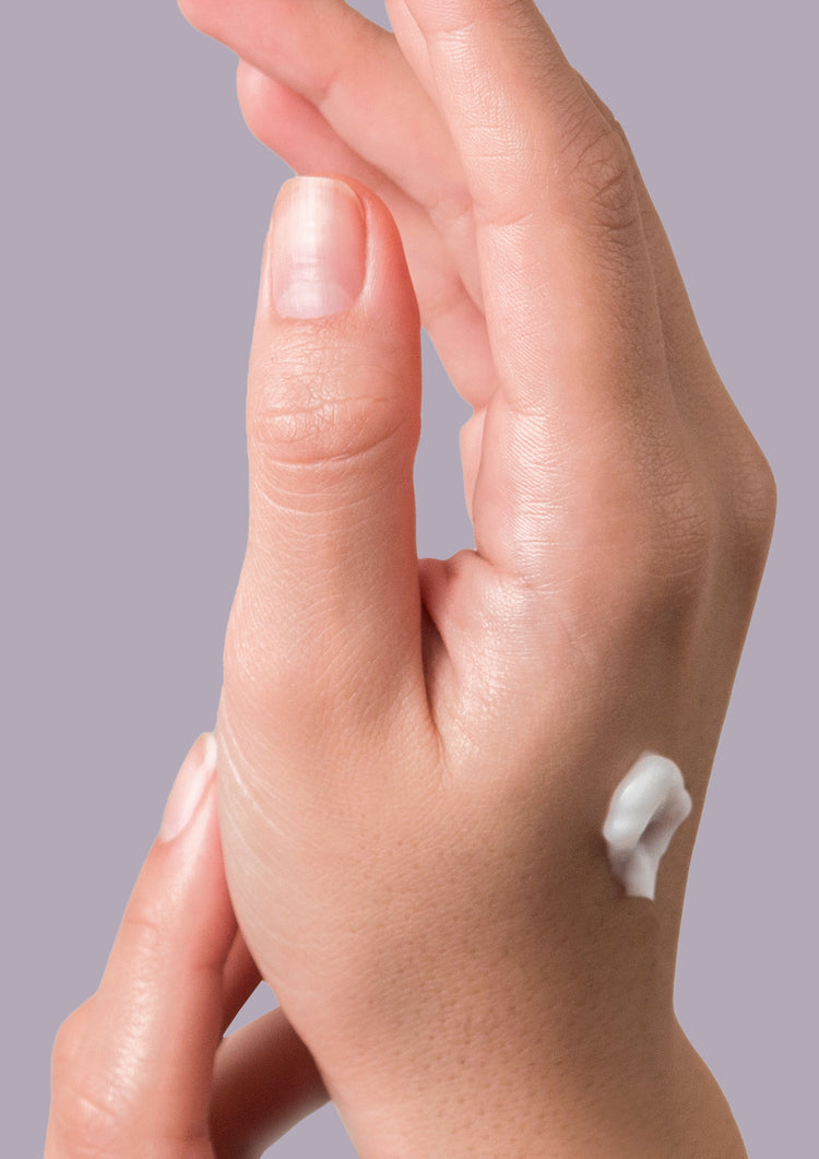 Hand with a dab of white hand cream on the back against a lilac grey background
