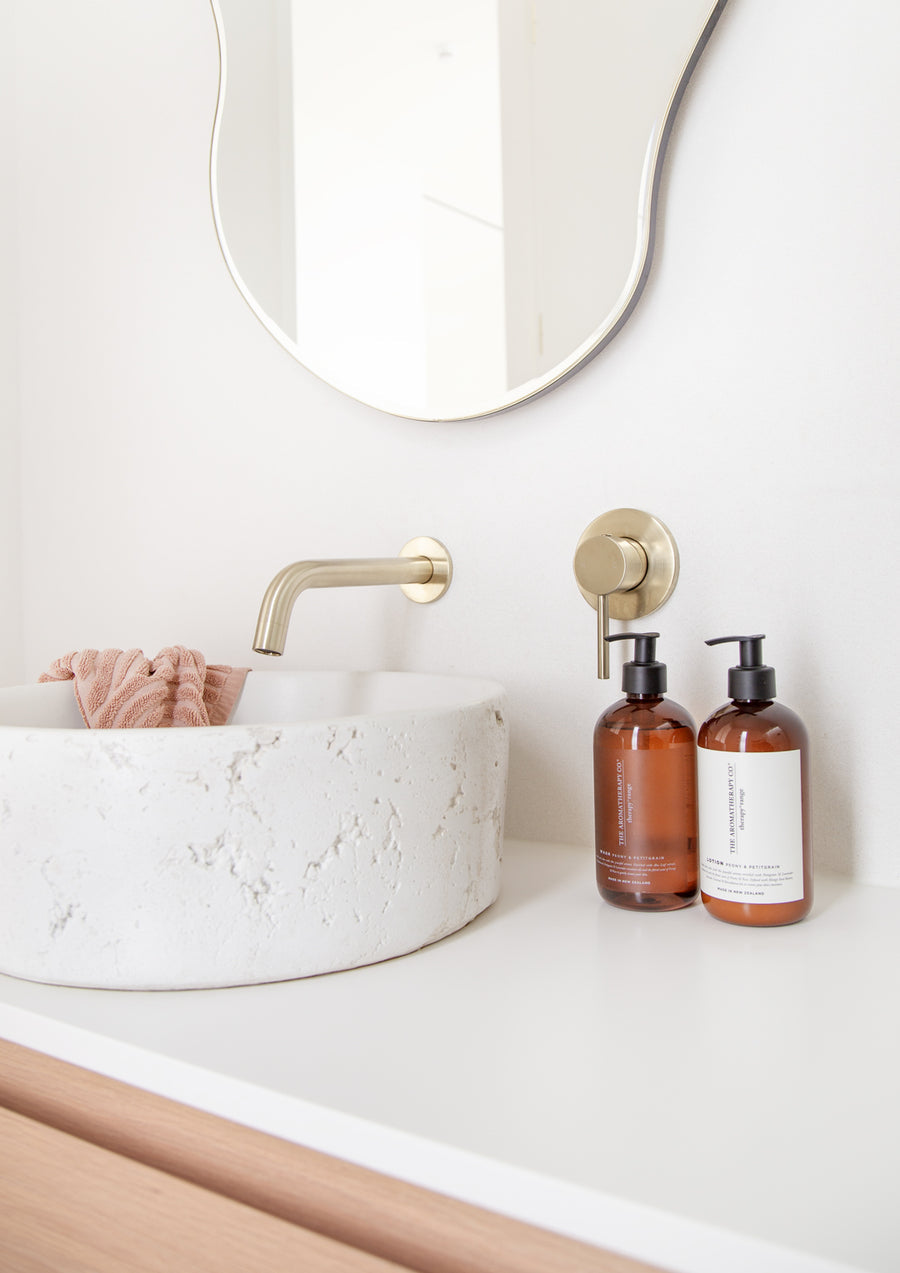 Two amber Therapy pump bottles on a white vanity beside a stone basin, brass tap and wavy mirror