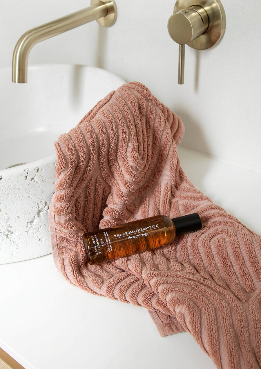 Amber room spray lying on a folded pink textured towel beside a white basin and brass taps