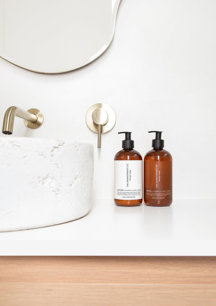 Therapy lotion and wash bottles on a white vanity beside a textured stone basin, brass tap and wavy mirror