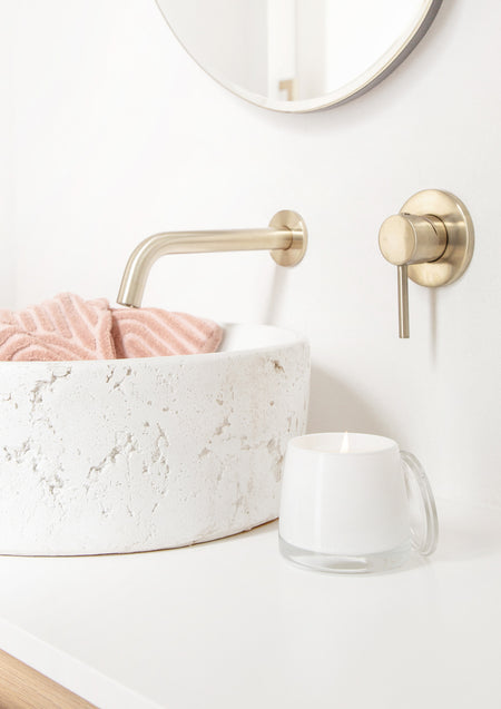 White glass candle on a vanity beside a stone basin with a pink towel and brass tapware