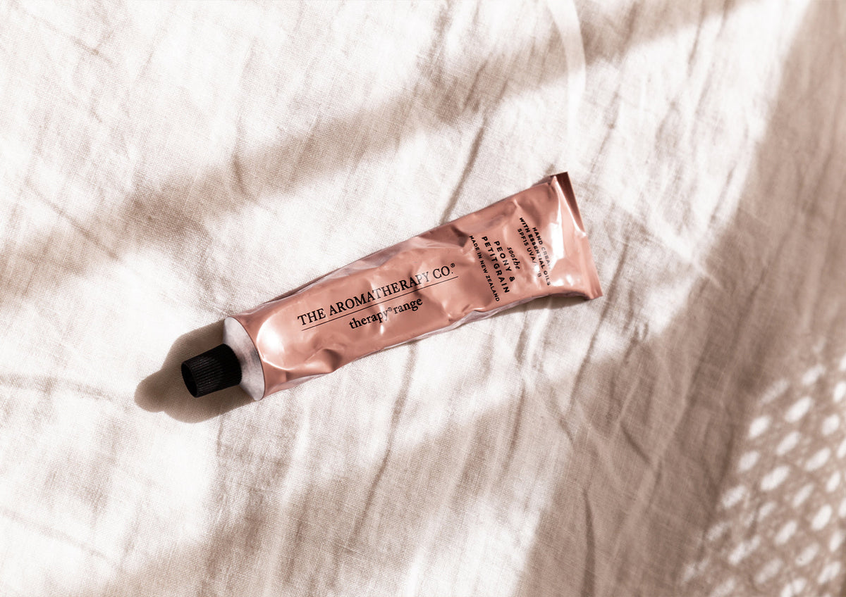 Blush hand cream tube lying across crumpled white linen in dappled light, landscape view