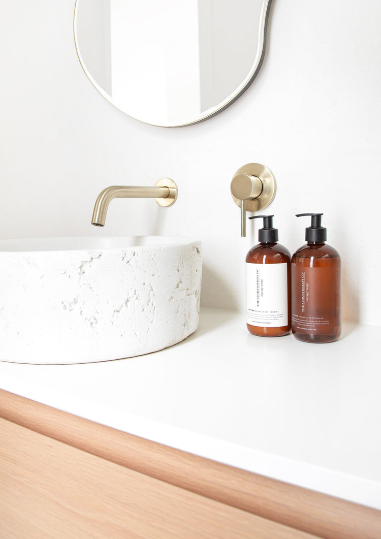 Therapy lotion and wash pump bottles beside a textured stone basin, brass tap and wavy mirror