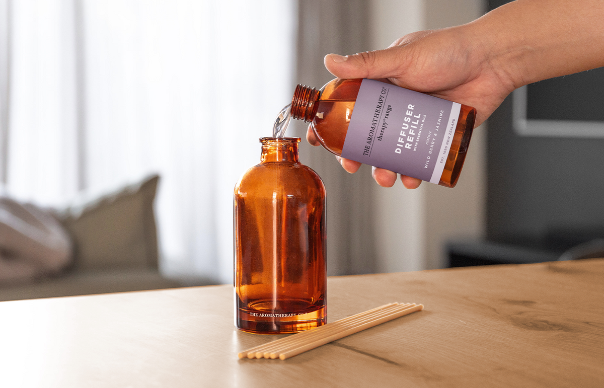 Hand pouring a Therapy diffuser refill into the amber bottle beside loose reeds