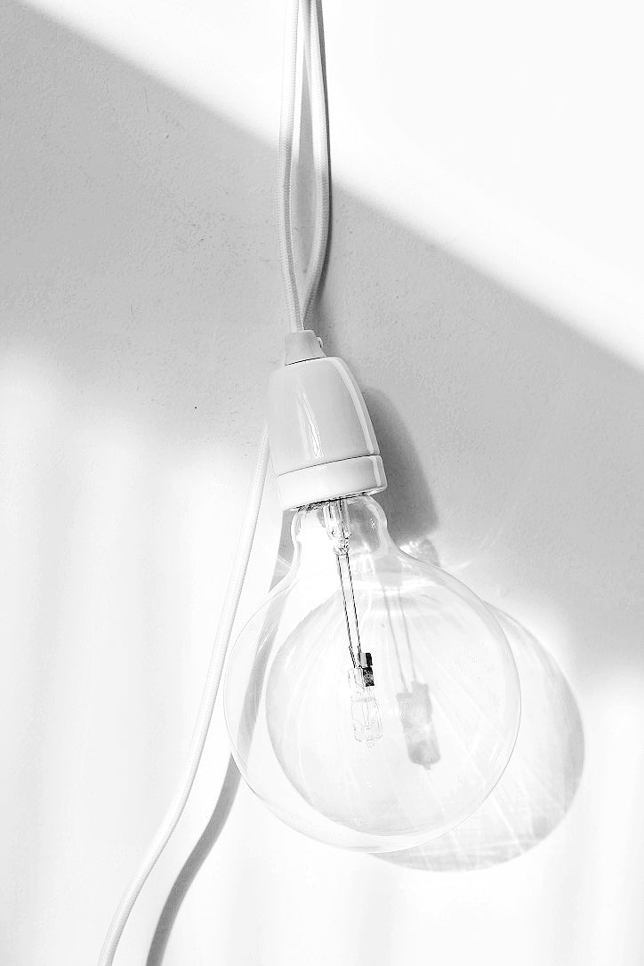 Minimalist clear bulb pendant light with white cord and fitting, hanging against a sunlit white wall.