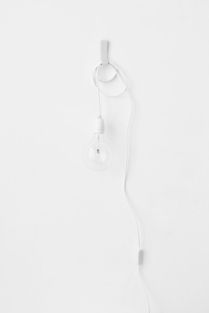 Minimalist plug-in pendant light with a clear bulb and white cord looped on a wall hook against a white wall.