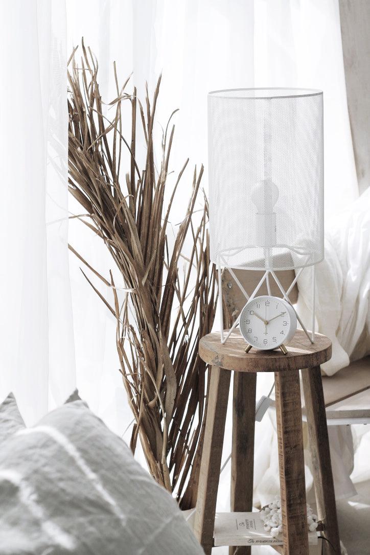White mesh cylinder lamp and a small clock on a rustic round timber stool beside dried grasses