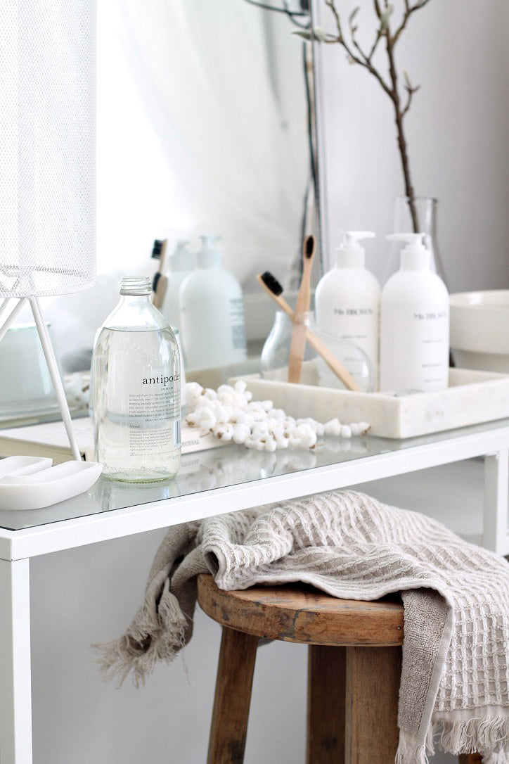 Rustic round timber stool holding a waffle linen throw beneath a white bathroom console with bottles