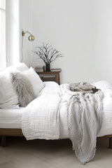 Bedroom styled with a white bed, layered grey linen throw, and soft pillows. A bedside table holds stacked books and a black sculptural branch arrangement.