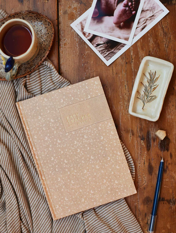 A Write To Me Baby Tracker Journal placed on a wooden table, alongside a cup of coffee.