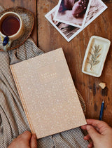 A person holding a notebook and a cup of tea on a wooden table, jotting down their baby's daily activities in the Write To Me Baby Tracker Journal.