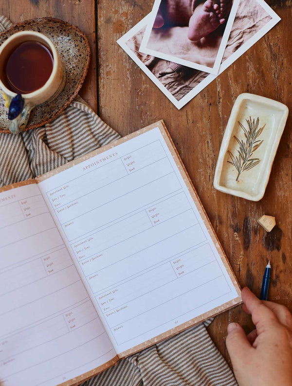 An open Write To Me Baby Tracker Journal with a cup of tea and a photo of a baby.