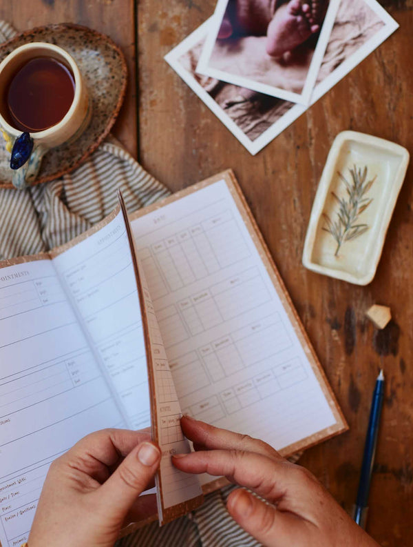 A woman's hand holding a Write To Me Baby Tracker Journal and a cup of tea on a wooden table.