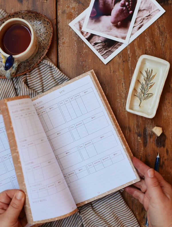 A hand holding a Write To Me Baby Tracker Journal and a cup of tea.