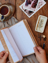 A woman's hands holding a Write To Me BABY TRACKER JOURNAL. OAT and a cup of coffee on a wooden table.