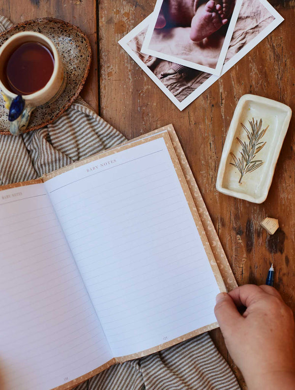 A woman holding a Write To Me Baby Tracker Journal - Oat and a cup of tea on a wooden table.