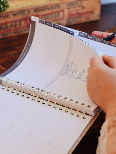A person diligently scheduling their monthly journey in a Write To Me 2024 Daily Hustle Planner, writing in a notebook on top of a wooden table.