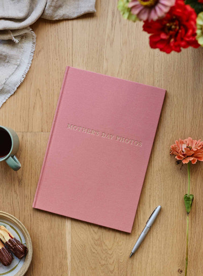 Blush linen photo journal closed on a timber table with flowers, a pen and a mug