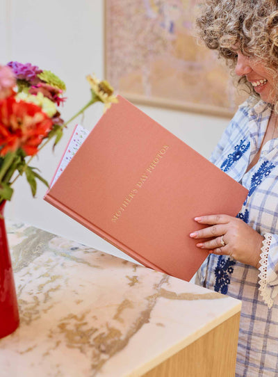 Person holding the blush linen photo journal upright beside a vase of flowers