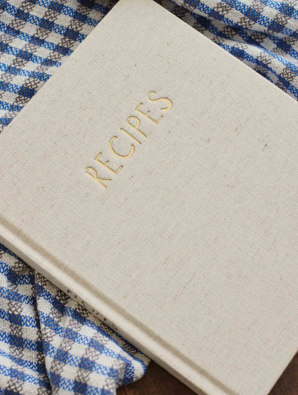 Oatmeal linen recipe journal cover with embossed title, angled on blue gingham