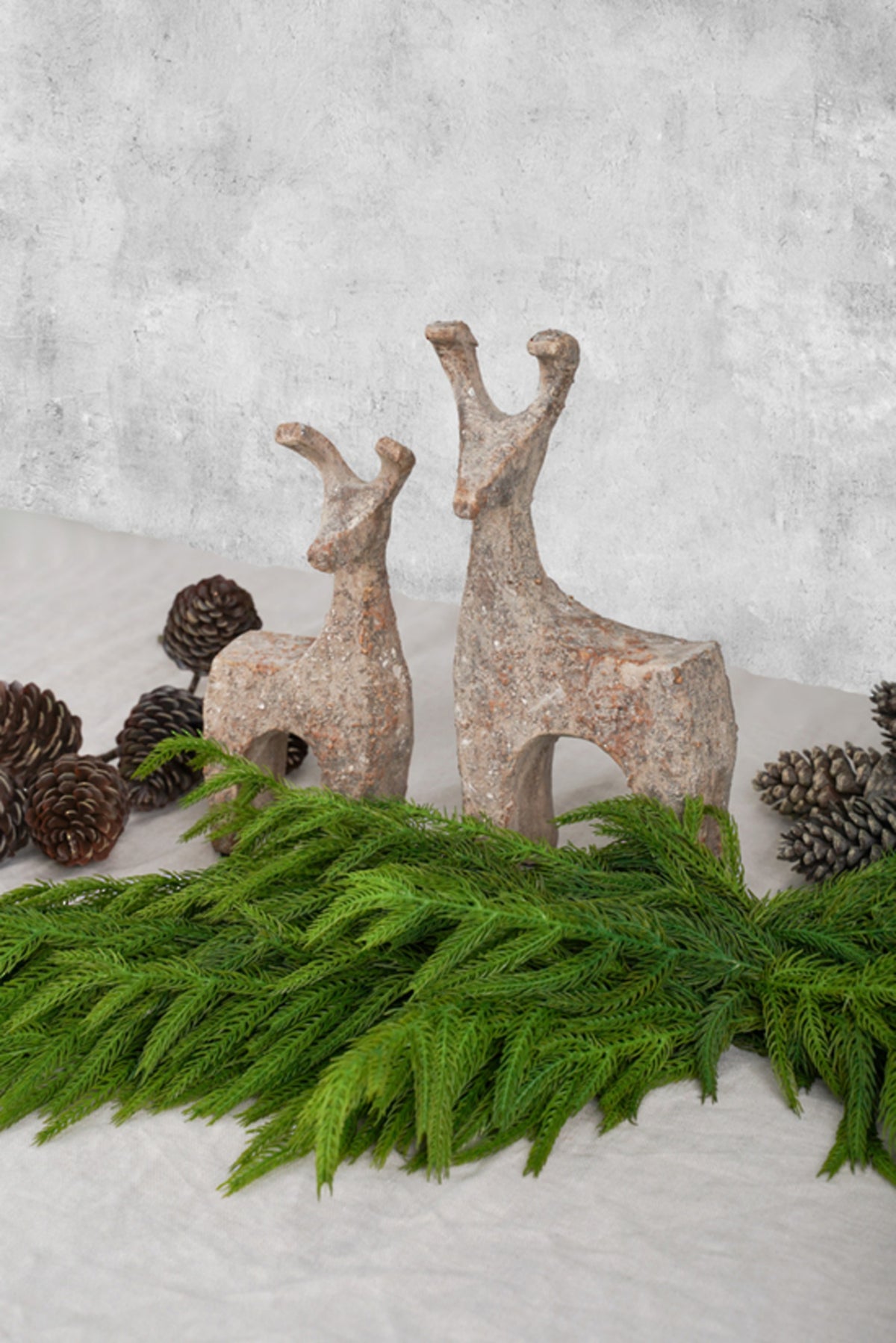 Two stone-look deer with a pine garland and pinecones on a grey concrete backdrop