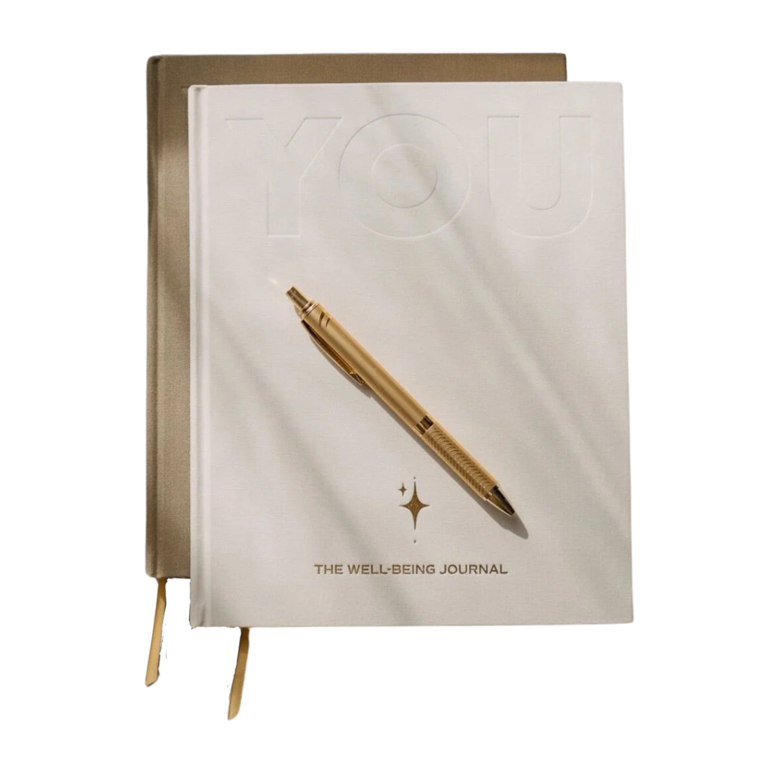 YOU. The Well-Being Journal in sand and cream, two copies stacked with a gold pen resting on the cover