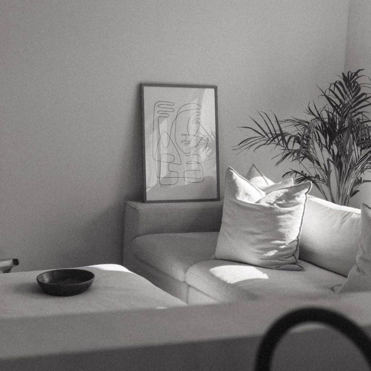 Abstract Faces print framed on a grey sofa beside cushions and a palm in a sunlit room