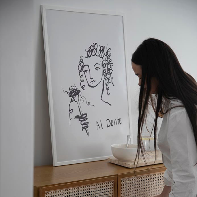 Al Dente print in a white frame on a rattan sideboard beside a woman and a pasta bowl