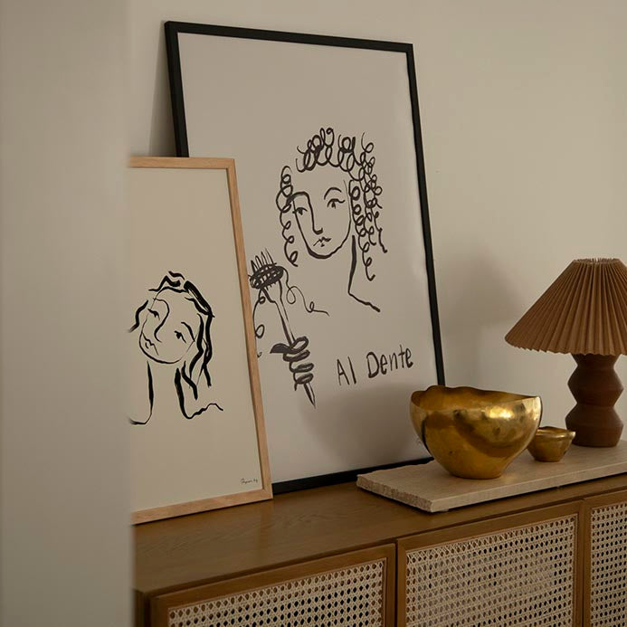 Al Dente print in a black frame on a rattan sideboard beside a gold bowl and pleated lamp