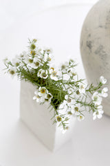 White Alba wax flower faux bush arranged in a white geometric ceramic vase beside a stone sphere on a neutral surface