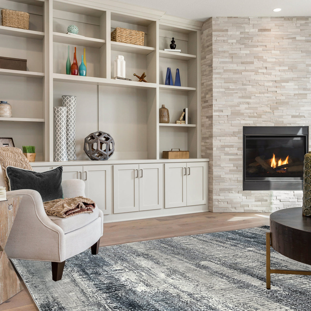 Blue and grey abstract rug in a lounge with white shelving, a stone fireplace and a cream armchair
