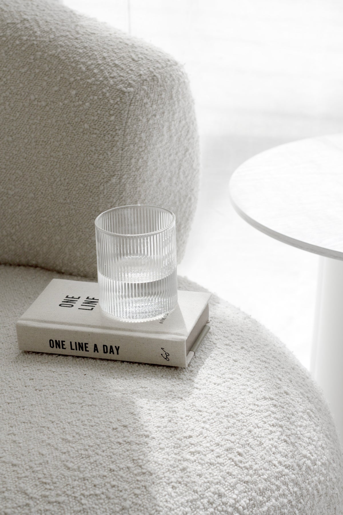 Fluted water glass on a journal resting on a cream boucle chair beside a white side table