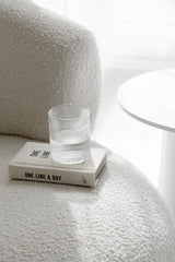 Clear fluted water glass resting on a One Line a Day journal on a boucle cream chair beside a white round side table in soft light.