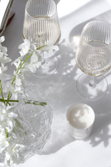 Two fluted wine glasses styled with white florals, a candle, and glass bowl in soft sunlight for a coastal, modern tablescape.