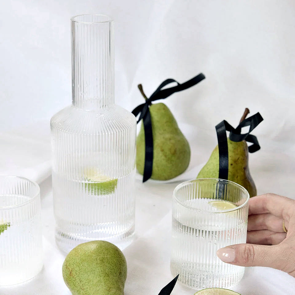 Hand holding a fluted glass beside the Amalfi decanter and green pears tied with black ribbon