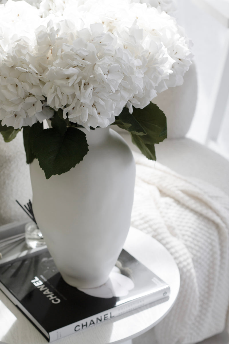 Artificial white hydrangea stems in matte sculptural vase styled on white side table with Chanel book and boucle chair – soft luxe look
