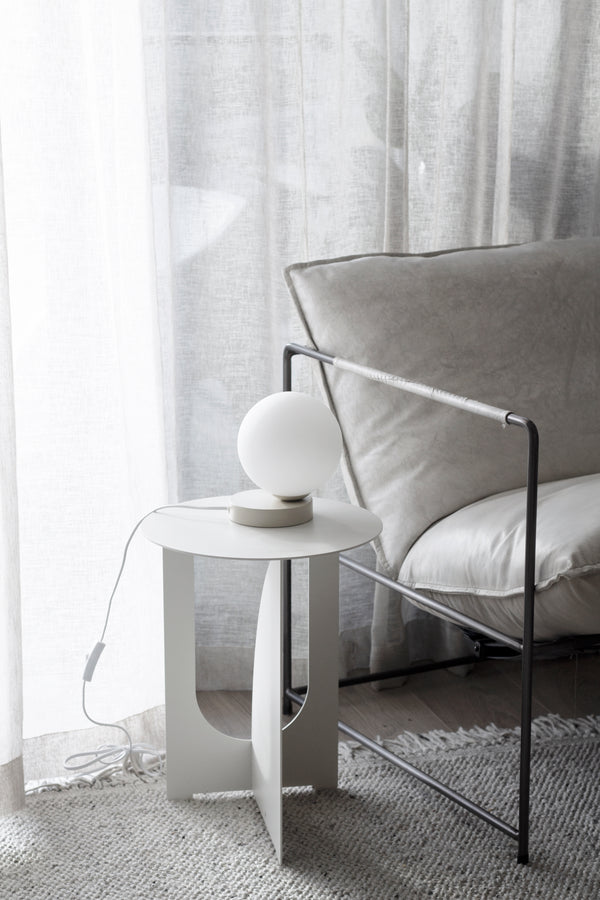 White Arch Side Table styled with a round lamp beside a grey armchair and sheer linen curtains.
