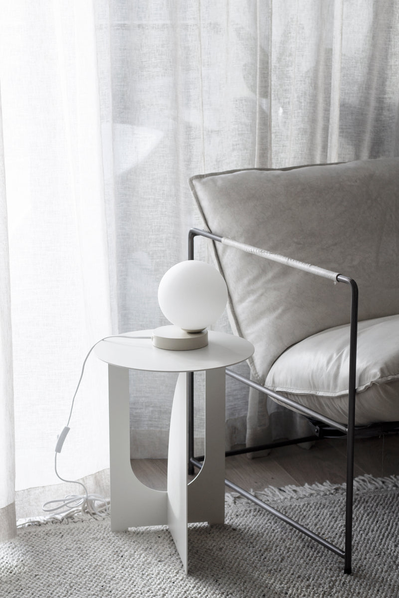 White Arch Side Table styled with a round lamp beside a grey armchair and sheer linen curtains.