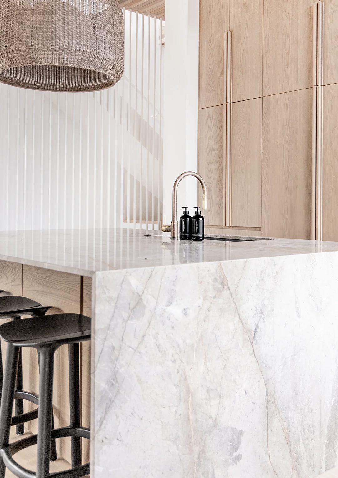Two black The Aromatherapy Co pump bottles beside a brass tap on a marble kitchen island, with oak cabinetry, black bar stools and a woven pendant light