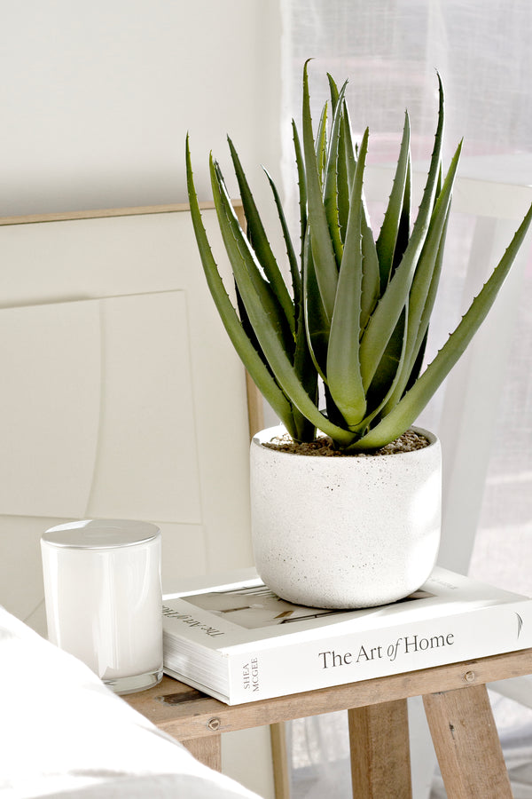 Artificial aloe plant in white cement pot styled on book and stool beside candle; realistic faux greenery for home decor.