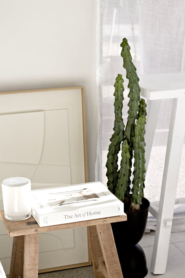Lifelike tall artificial cactus in black pot styled beside books, natural wood stool, and framed neutral artwork in a minimalist interior.