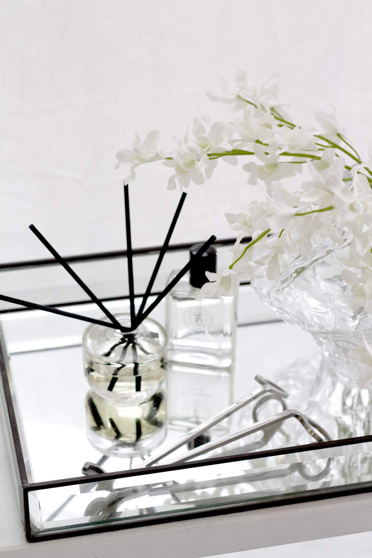 White dendrobium orchid faux flower bundle styled in a clear textured glass vase on a mirrored tray with a reed diffuser, perfume bottle and wick trimmer