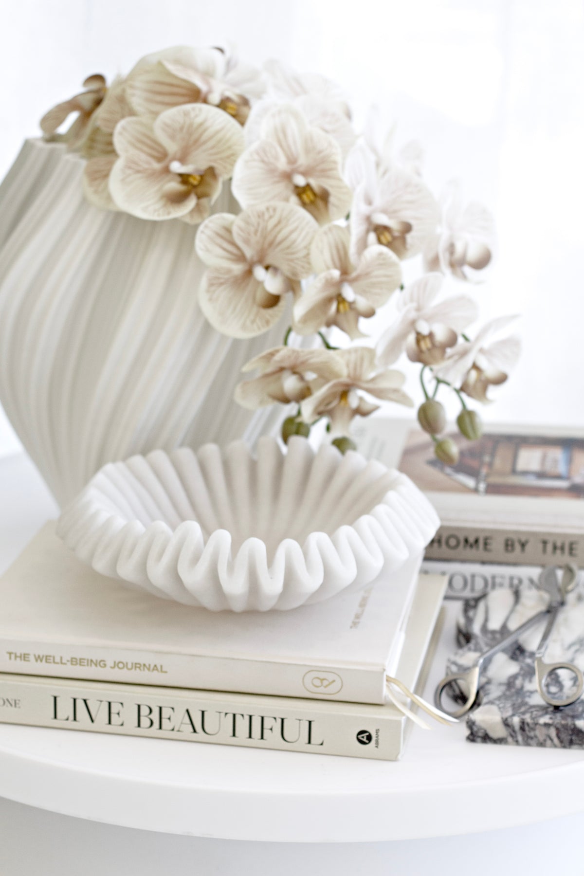 Orchids in a ribbed white vase on a round table beside a fluted dish and stacked books