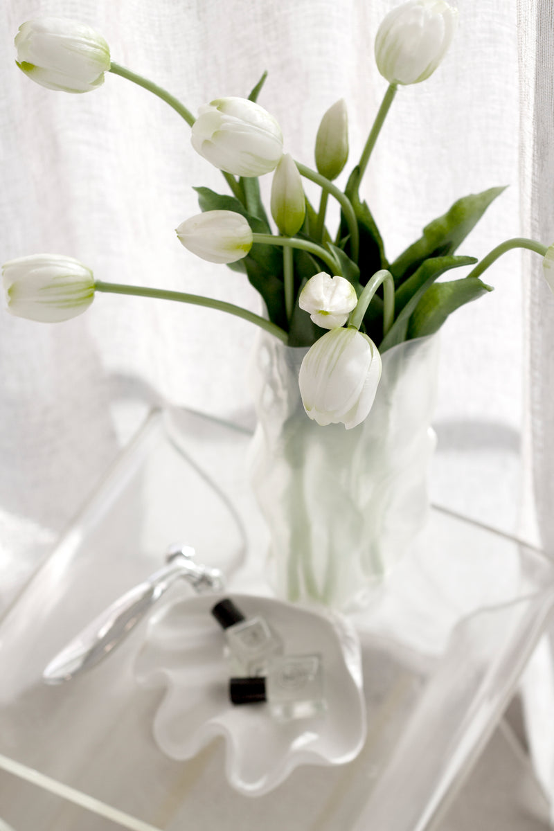 Artificial white tulip bouquet styled in a frosted glass vase on a clear table with soft curtains.