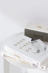 Atomic Habits by James Clear styled on a white side table with Good Vibes, Good Life, a clear perfume bottle, and a cream Marshall speaker.