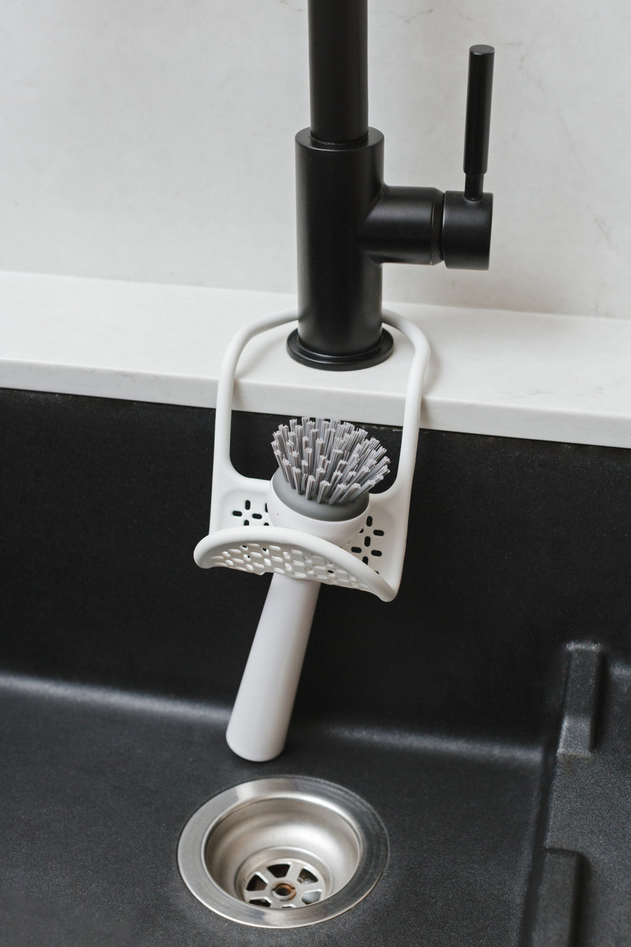 Umbra Sling sink caddy in white slung over a black kitchen tap, holding a scrubbing brush