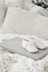 Soft grey linen baby journal titled “BABY – The First Five Years” resting on a chunky white knit blanket with tiny white baby moccasins on top and textured cream cushions in the background.